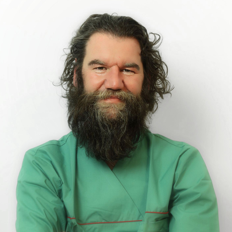 Un uomo con barba lunga e riccia e capelli ondulati indossa un camice medico verde e sorride dolcemente, su uno sfondo bianco.