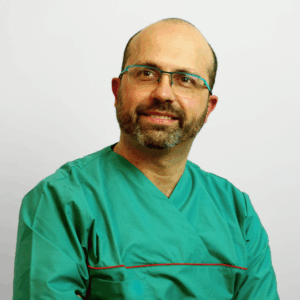 Un uomo di mezza età con occhiali e barba indossa un camice medico verde e sorride leggermente guardando di lato, in posa su un semplice sfondo bianco.