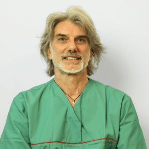 Un uomo di mezza età con capelli e barba grigi sorride e indossa un camice medico verde su uno sfondo bianco.