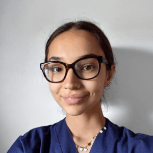 Una giovane donna dalla pelle marrone chiaro, con grandi occhiali neri e un top blu navy, sorride dolcemente alla telecamera. Ha i capelli lisci e castani legati all'indietro e indossa una collana con piccoli ciondoli.