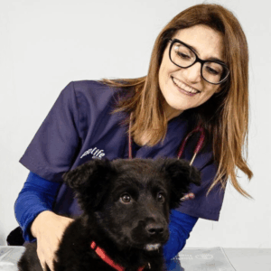 Un veterinario sorridente con occhiali e camice esamina un cucciolo nero con un collare rosso. La veterinaria ha uno stetoscopio al collo e tiene delicatamente il cucciolo su un tavolo da visita.