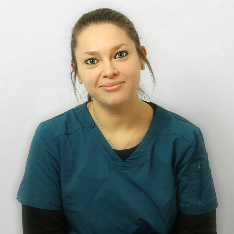 Una donna dalla pelle chiara e dai capelli castani legati all'indietro indossa un camice medico verde acqua su una camicia nera e sorride dolcemente mentre è seduta su uno sfondo chiaro.